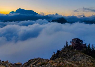 Clouds float below a fire lookout with mountains and a sunrise in the background.
