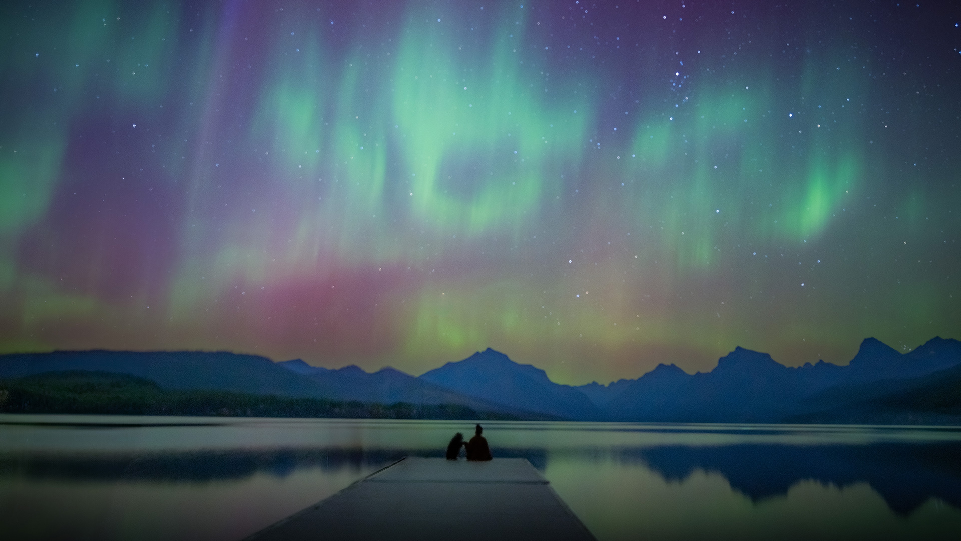 A person sitting on a dock watching the northern lights above a lake and mountains.
