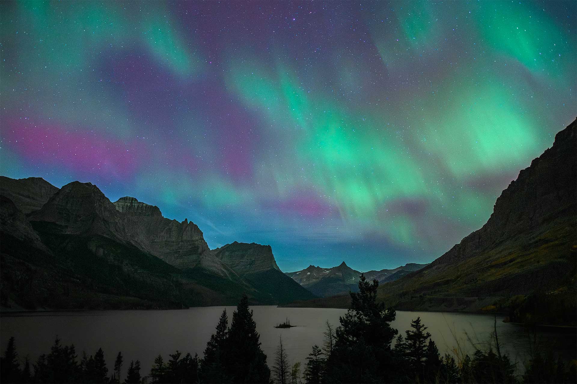 Green and purple northern lights fill the sky above mountains and a lake with a small island in the center.