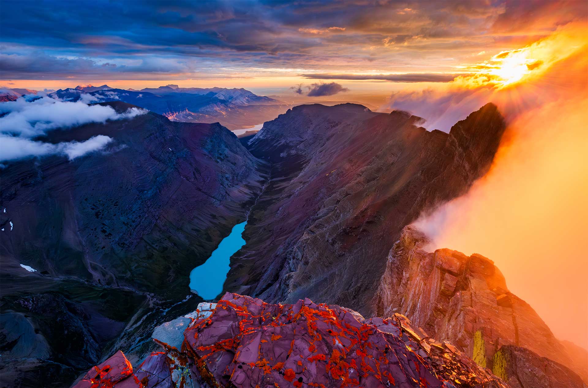 A blue lake below a mountain ridge with orange clouds on the right side and the sun rising.