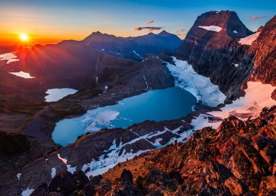 A mountain lake with a glacier surrounded by mountains and the sun rising in the background.