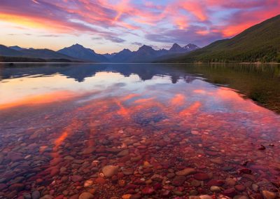 Pink clouds and mountains reflecting on a lake with red rocks below the surface.